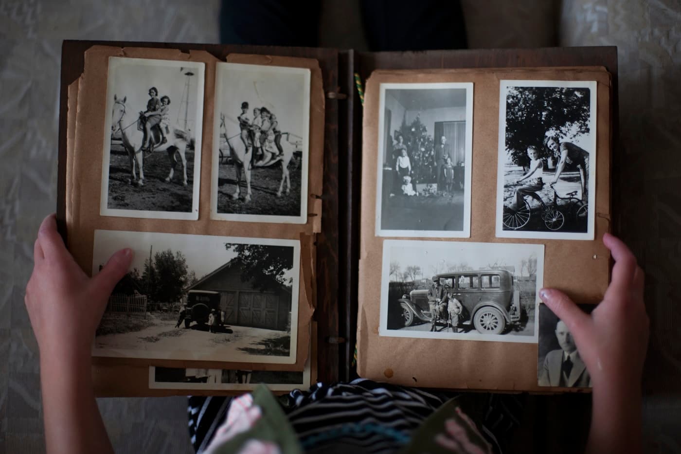 Hands holding open a vintage family photo album filled with old black-and-white photographs — childhood, horses, a classic car, a bicycle — evoking a lifetime of stories worth preserving